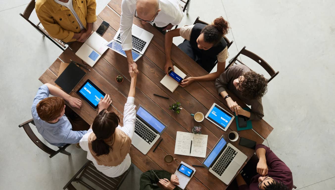 |business-meeting-around-a-table-2-people-shaking-hands-laptops-phones-on-table
