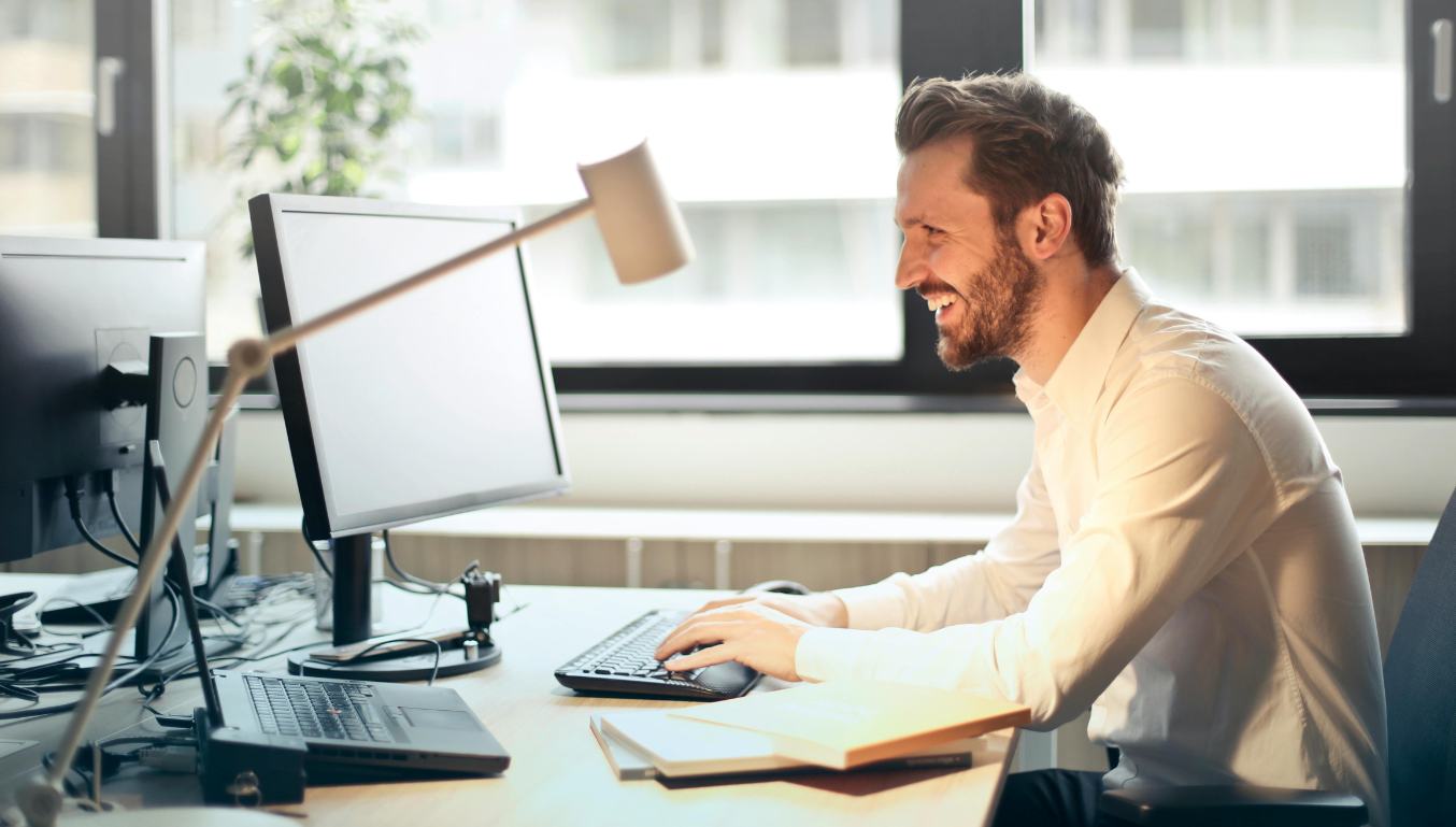 Man in White Dress Shirt Sitting on Black Rolling Chair While Facing Black Computer Set and Smiling