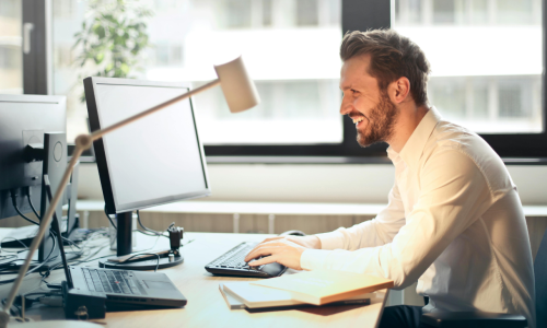 Man in White Dress Shirt Sitting on Black Rolling Chair While Facing Black Computer Set and Smiling